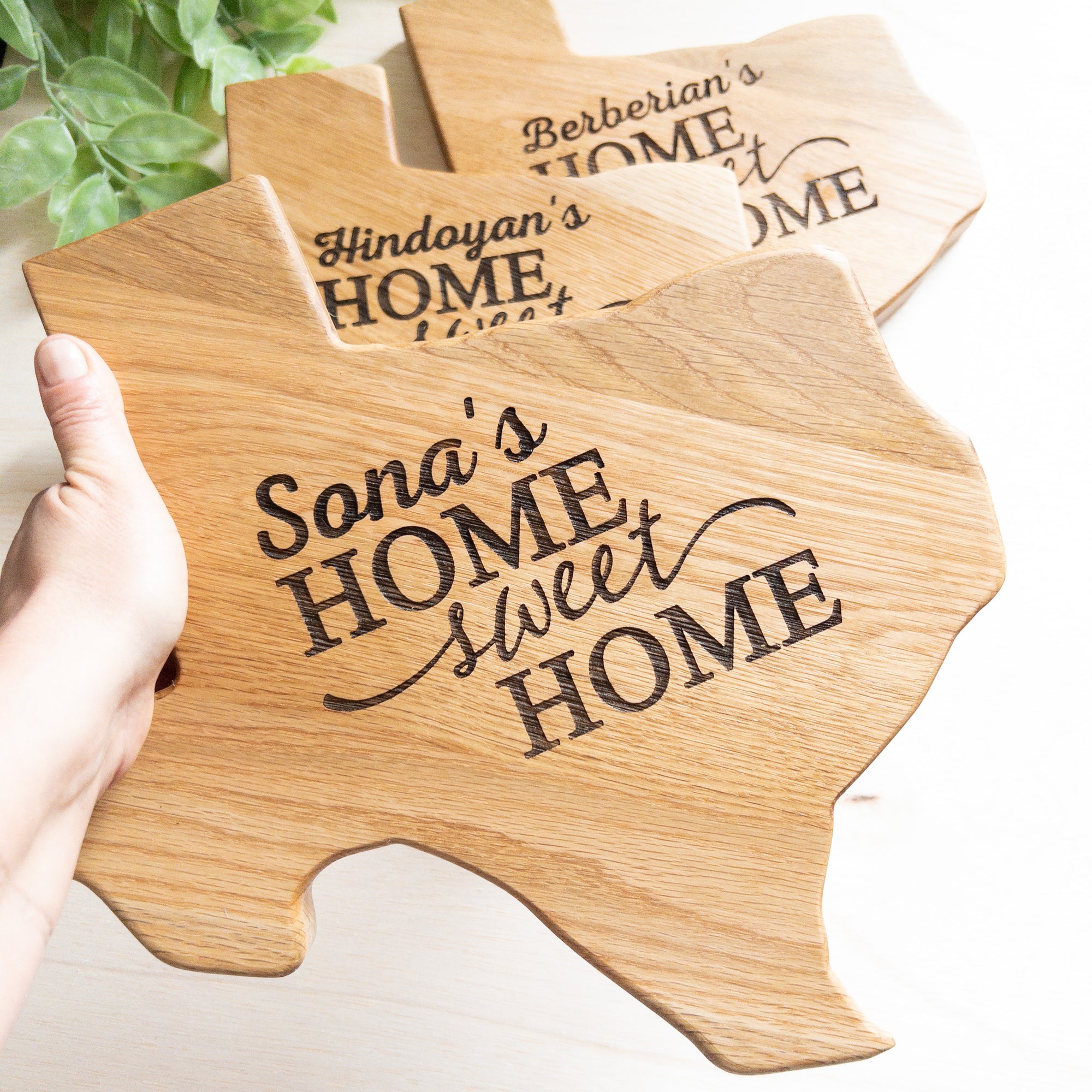 An overhead shot of a rectangular wooden chopping board with a detailed handcrafted design of the state of Texas, including its borders and major cities. The board is surrounded by kitchen utensils and ingredients, and is placed on a countertop. The wood grain is visible and the surface of the board looks smooth and polished.